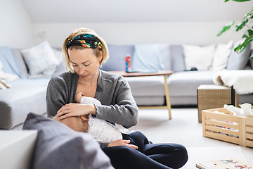 Image showing Young woman breastfeeding her infant baby boy casualy sitting on child's playing mat on living room floor at home.