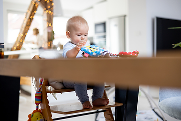 Image showing Happy infant sitting at dining table and playing with his toy in traditional scandinavian designer wooden high chair in modern bright atic home. Cute baby playing with toys