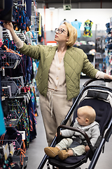 Image showing Casualy dressed mother choosing sporty shoes and clothes products in sports department of supermarket store with her infant baby boy child in stroller.