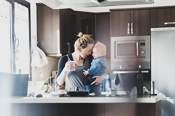 Image showing Happy mother holding her little infant baby boy while drinking morning coffee and making pancakes for breakfast in domestic kitchen. Family lifestyle, domestic life concept.