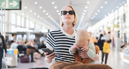 Image showing Mother traveling with child, holding his infant baby boy at airport terminal, checking flight schedule, waiting to board a plane. Travel with kids concept.