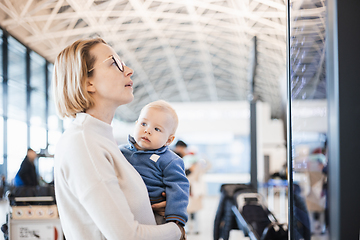 Image showing Mother traveling with child, holding his infant baby boy at airport terminal, checking flight schedule, waiting to board a plane. Travel with kids concept.