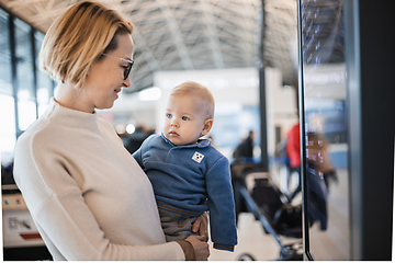 Image showing Mother traveling with child, holding his infant baby boy at airport terminal, checking flight schedule, waiting to board a plane. Travel with kids concept.