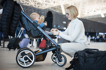 Image showing Motherat interacting with her infant baby boy child in stroller while travelling at airport terminal station. Travel with child concept.