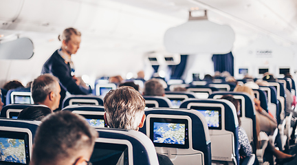 Image showing Interior of airplane with passengers on seats and stewardess in uniform walking the aisle, serving people. Commercial economy flight service concept.