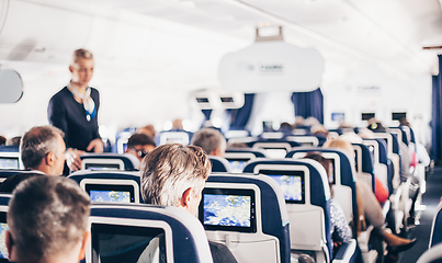 Image showing Interior of airplane with passengers on seats and stewardess in uniform walking the aisle, serving people. Commercial economy flight service concept.