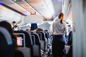 Image showing Interior of airplane with passengers on seats and stewardess in uniform walking the aisle, serving people. Commercial economy flight service concept.