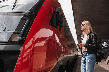 Image showing Young blond woman in jeans, shirt and leather jacket wearing bag and sunglass, embarking red modern speed train on train station platform. Travel and transportation.