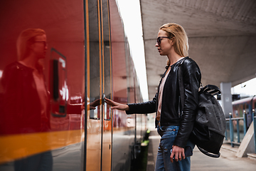 Image showing Young blond woman in jeans, shirt and leather jacket wearing bag and sunglass, presses door button of modern speed train to embark on train station platform. Travel and transportation.