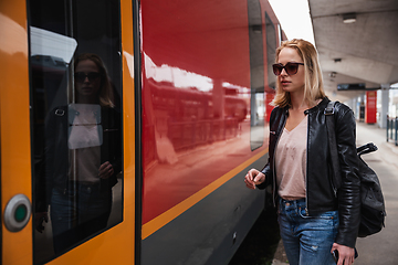 Image showing Young blond woman in jeans, shirt and leather jacket wearing bag and sunglass, embarking red modern speed train on train station platform. Travel and transportation.