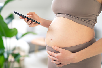 Image showing Close-up photo of pregnant female belly. Woman holding and using mobile smart phone application at home interiors. Pregnancy, technology, online shopping, preparation and expectation concept.