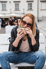 Image showing Pretty young blonde funny woman eating hamburger outdoor on the street.