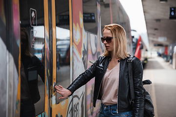 Image showing Young blond woman in jeans, shirt and leather jacket wearing bag and sunglass, presses door button of modern speed train to embark on train station platform. Travel and transportation.
