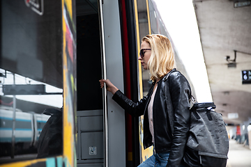 Image showing Young blond woman in jeans, shirt and leather jacket wearing bag and sunglass, presses door button of modern speed train to embark on train station platform. Travel and transportation.