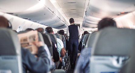 Image showing Interior of airplane with passengers on seats and stewardess in uniform walking the aisle, serving people. Commercial economy flight service concept.