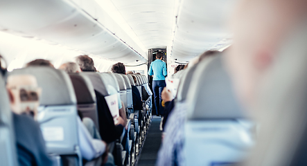 Image showing Interior of airplane with passengers on seats and stewardess in uniform walking the aisle, serving people. Commercial economy flight service concept.