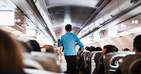 Image showing Interior of airplane with passengers on seats and stewardess in uniform walking the aisle, serving people. Commercial economy flight service concept.