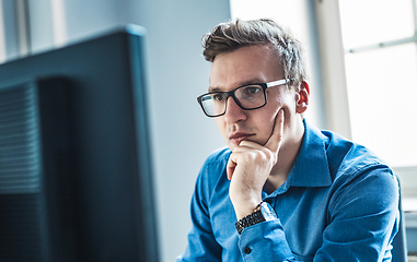 Image showing Handsome Young Businessman Wearing Eyeglasses Sitting at his Table Inside the Office, Looking at the Report on his Computer Screen.