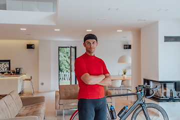 Image showing Photo of a determined triathlete standing in a modern large living room, behind him is his training bike
