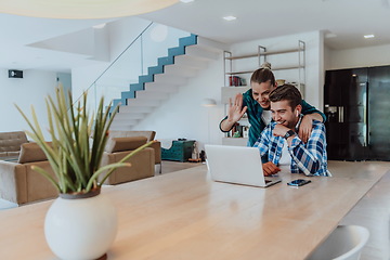 Image showing A young married couple is talking to parents, family and friends on a video call via a laptop while sitting in the living room of their modern house