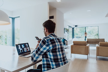 Image showing The man sitting at a table in a modern living room, using a smartphone and laptop for business video chat, conversation with friends and entertainment