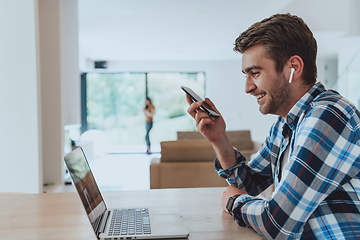 Image showing The man sitting at a table in a modern living room, using a smartphone and laptop for business video chat, conversation with friends and entertainment