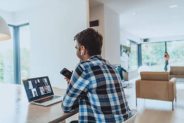 Image showing The man sitting at a table in a modern living room, using a smartphone and laptop for business video chat, conversation with friends and entertainment