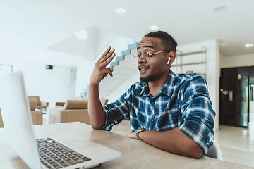 Image showing African American man in glasses sitting at a table in a modern living room, using a laptop for business video chat, conversation with friends and entertainment