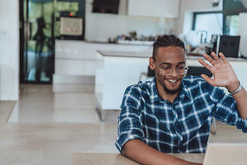 Image showing African American man in glasses sitting at a table in a modern living room, using a laptop for business video chat, conversation with friends and entertainment