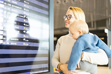 Image showing Mother traveling with child, holding his infant baby boy at airport terminal, checking flight schedule, waiting to board a plane. Travel with kids concept.