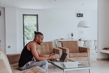 Image showing African American sport man in glasses sitting at a table in a modern living room, using a laptop for business video chat, conversation with friends and entertainment