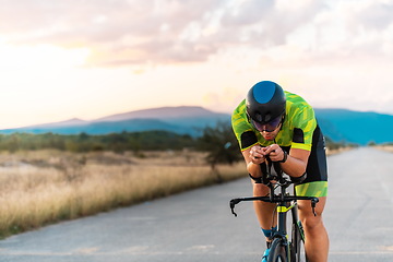 Image showing Triathlete riding his bicycle during sunset, preparing for a marathon. The warm colors of the sky provide a beautiful backdrop for his determined and focused effort.