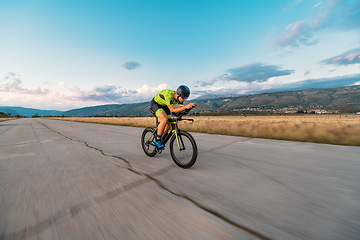 Image showing Triathlete riding his bicycle during sunset, preparing for a marathon. The warm colors of the sky provide a beautiful backdrop for his determined and focused effort.