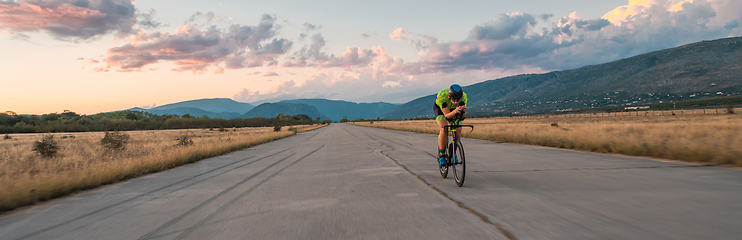 Image showing Triathlete riding his bicycle during sunset, preparing for a marathon. The warm colors of the sky provide a beautiful backdrop for his determined and focused effort.