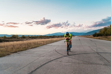 Image showing Triathlete riding his bicycle during sunset, preparing for a marathon. The warm colors of the sky provide a beautiful backdrop for his determined and focused effort.