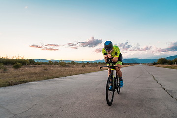 Image showing Triathlete riding his bicycle during sunset, preparing for a marathon. The warm colors of the sky provide a beautiful backdrop for his determined and focused effort.