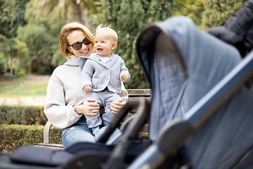 Image showing Mother sitting on bench in urban park, laughing cheerfully, holding her smiling infant baby boy child in her lap having baby stroller parked by their site.