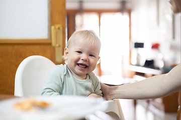 Image showing Adorable cheerful happy infant baby boy child smiling while sitting in high chair at the dining table in kitchen at home beeing spoon fed by his mother