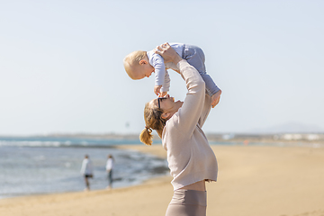 Image showing Mother enjoying summer vacations holding, playing and lifting his infant baby boy son high in the air on sandy beach on Lanzarote island, Spain. Family travel and vacations concept.