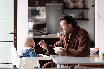 Image showing Father wearing bathrope spoon feeding hir infant baby boy child sitting in high chair at the dining table in kitchen at home in the morning.