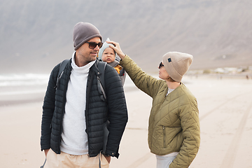 Image showing Young father carrying his infant baby boy son in backpack on windy sandy beach. Family travel and winter vacation concept.