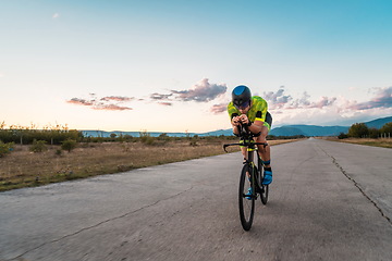 Image showing Triathlete riding his bicycle during sunset, preparing for a marathon. The warm colors of the sky provide a beautiful backdrop for his determined and focused effort.