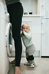 Image showing Little infant baby boy child hiding between mothers legs demanding her attention while she is multitasking, trying to do some household chores in kitchen at home. Mother on maternity leave.