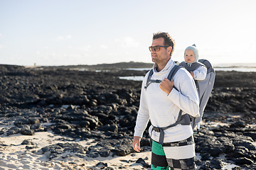 Image showing Young father carrying his infant baby boy son in backpack on black rock volcanic beach on Lanzarote island, Spain. Family travel and winter vacation concept.