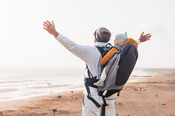 Image showing Young father rising hands to the sky while enjoying pure nature carrying his infant baby boy son in backpack on windy sandy beach. Family travel concept.