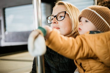 Image showing Mother carries her child while standing and holding on to the bus. Mom holding her infant baby boy in her arms while riding in a public transportation. Cute toddler boy traveling with his mother.