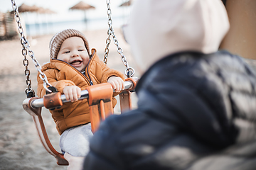 Image showing Mother pushing her cheerful infant baby boy child on a swing on sandy beach playground outdoors on nice sunny cold winter day in Malaga, Spain.