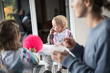 Image showing Mother spoon feeding her baby boy child in baby high chair with fruit puree at dinning table at home. Baby solid food introduction concept.