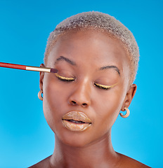 Image showing Face, makeup and eyeshadow with a black woman on a blue background in studio to apply a beauty product. Cosmetics, gold color on a brush and a young female model eyes closed for style or glamour