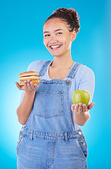 Image showing Portrait, smile and woman with burger, apple and choice in studio isolated on a blue background. Face, fruit and person with sandwich, fast food or decision for healthy diet, nutrition or wellness.